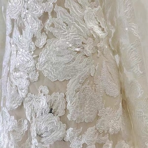 May include: White floral lace fabric with sequins. The fabric has a delicate, intricate design with a floral pattern. The lace is sheer and has a soft, flowing texture.