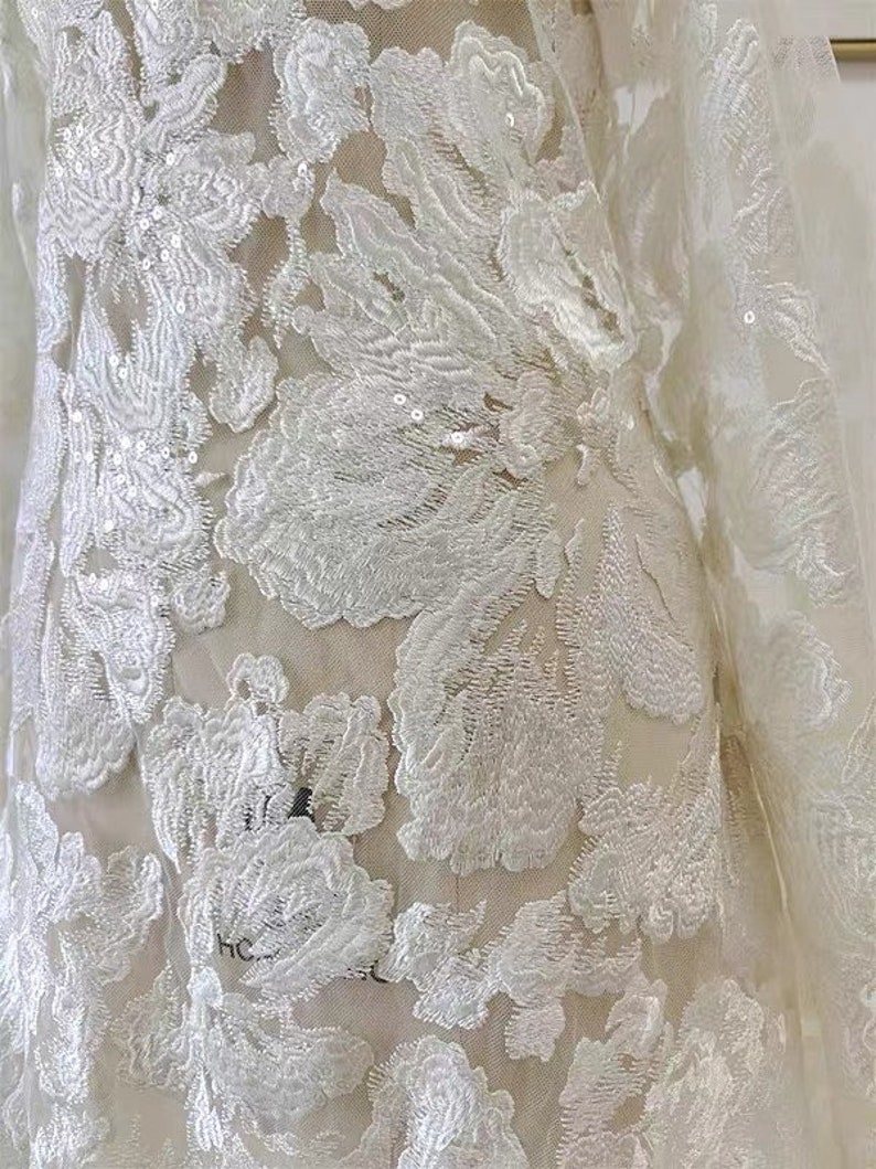 May include: White floral lace fabric with sequins. The fabric has a delicate, intricate design with a floral pattern. The lace is sheer and has a soft, flowing texture.