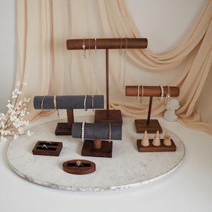 May include: A collection of jewelry display stands made of dark wood and gray velvet. The stands hold necklaces, bracelets, and rings. The jewelry is gold and silver, and the background is a neutral beige.