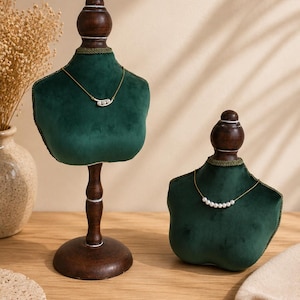 May include: Two dark green velvet jewelry display busts with wooden stands. The taller bust features a gold chain necklace with three small white pearls. The shorter bust displays a gold chain necklace with a row of six small white pearls.