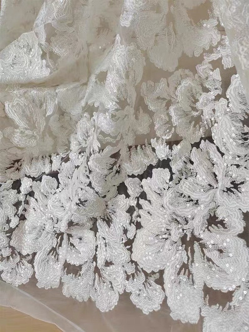 May include: White floral lace fabric with sequins. The lace has a delicate floral pattern and is made of a sheer material. The sequins are small and scattered throughout the fabric.
