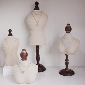May include: Four beige jewelry display busts with dark brown wooden stands. Each bust displays a gold necklace. One necklace has a single pearl, another has a series of pearls, and the other two have a small pendant. The word "PARIS" is printed on each bust.