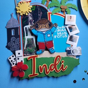 May include: A colorful, layered paper craft with a blue background. It features a cartoon frog character, palm trees, a Puerto Rican flag, and the word "Indi". Additional elements include photos, a camera, and dominoes.