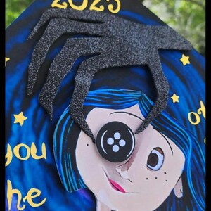 May include: A graduation cap featuring a cartoon illustration of a character with blue hair and a button eye. The cap has the year 2025 and a black spider-like design. The background is a starry night sky with text.