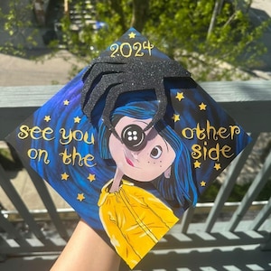 May include: A decorated graduation cap featuring a character with a button eye, blue hair, and a yellow jacket. The cap has the text "See you on the other side" and "2024" with star accents. A black spider-like embellishment is on top.