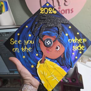 May include: A graduation cap featuring a Coraline-inspired design. The cap has a dark blue background with a starry night sky and the words "See you on the other side." The design includes a character with a button eye and the year 2026.