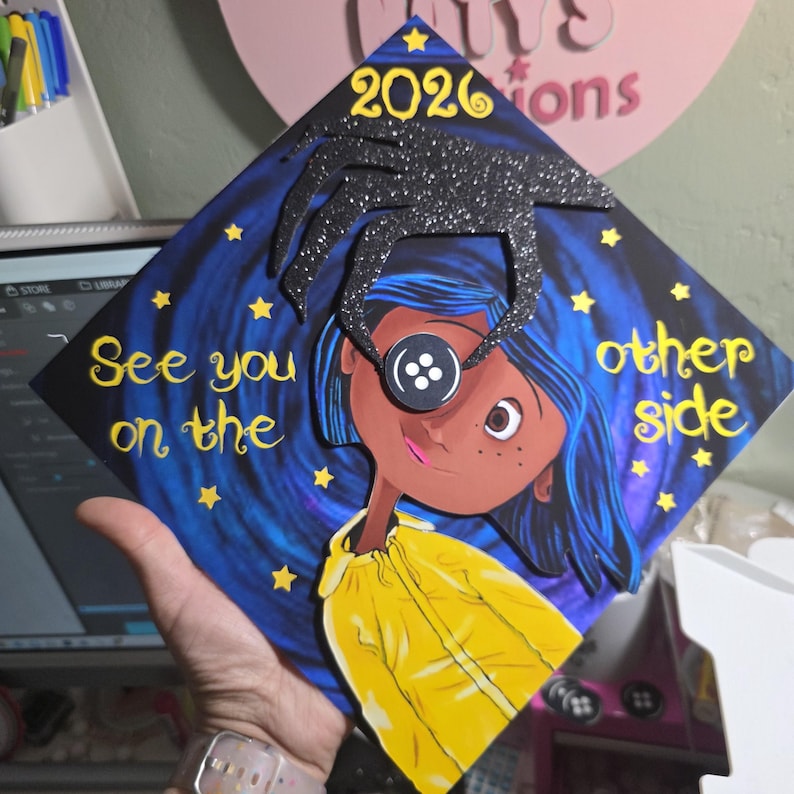 May include: A graduation cap featuring a Coraline-inspired design. The cap has a dark blue background with a starry night sky and the words "See you on the other side." The design includes a character with a button eye and the year 2026.