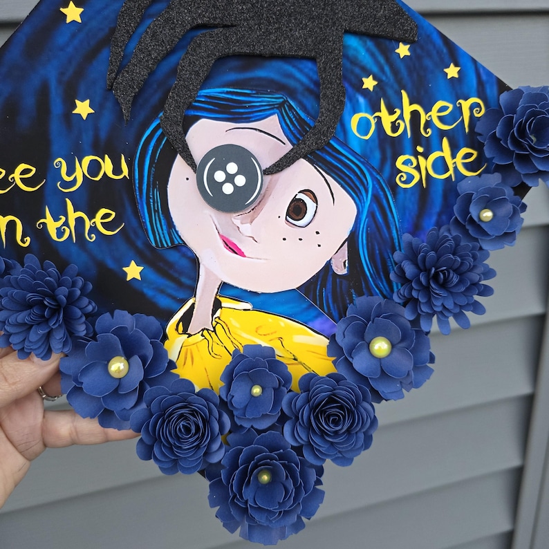 May include: A decorated graduation cap featuring a cartoon illustration of a girl with a button eye, surrounded by blue paper flowers. The cap has the words "see you on the other side" in yellow script, set against a dark blue starry background.
