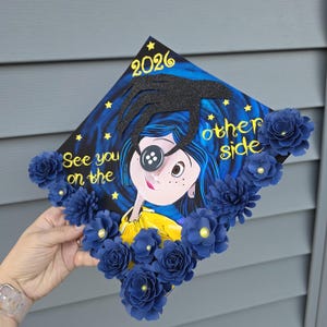 May include: A decorated graduation cap featuring a cartoon character with a button eye, surrounded by blue paper flowers. The cap has the text "See you on the other side" and the year "2026" in gold lettering.