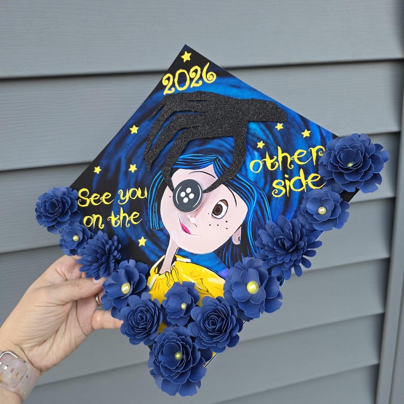 May include: A decorated graduation cap featuring a cartoon character with a button eye, surrounded by blue paper flowers. The cap has the text "See you on the other side" and the year "2026" in gold lettering.