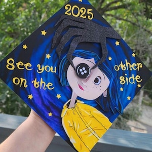 May include: A graduation cap featuring a cartoon illustration with the text "See you on the other side" and the year "2025". The design includes a character with a button eye and a black hand reaching down.
