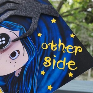 May include: A decorated graduation cap featuring a cartoon character with blue hair and a button eye. The cap has the text "2025" and "other side" in yellow, with yellow star accents against a blue and black background.