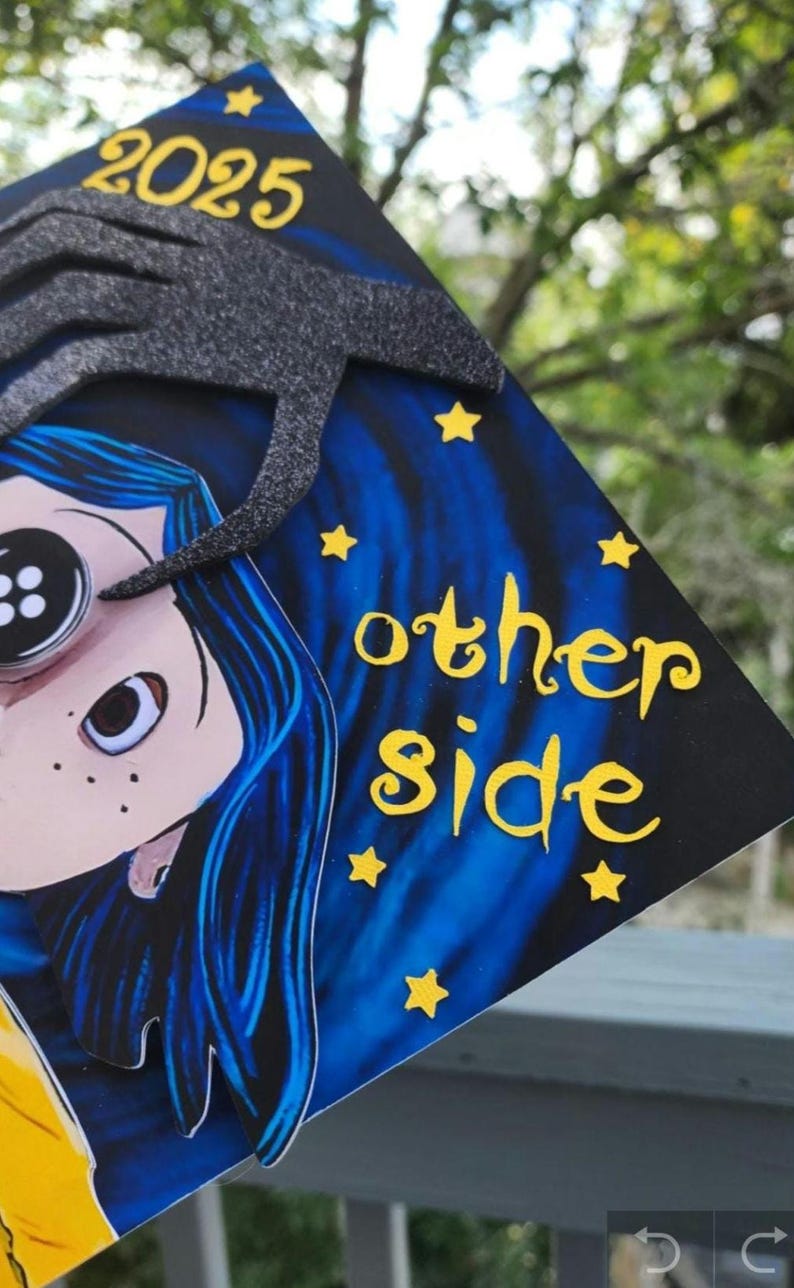 May include: A decorated graduation cap featuring a cartoon character with blue hair and a button eye. The cap has the text "2025" and "other side" in yellow, with yellow star accents against a blue and black background.