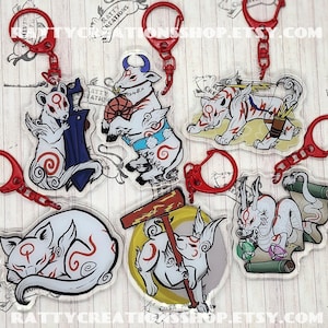 May include: A collection of acrylic keychains featuring white and red stylized animal characters with red key rings. The keychains have various designs, including a sleeping animal, a character with a hammer, and a character with a scroll. The text "RATTYCREATIONSSHOP.ETSY.COM" is visible.