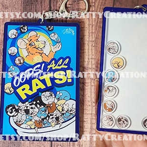 May include: A rectangular keychain with a blue border and a silver keyring. The front features a cartoon rat with the text "OOPS! ALL RATS!". The back has a white surface with several small, round rat-themed charms.