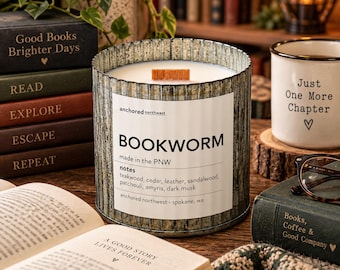 Puede incluir: Una vela cilíndrica en un recipiente de metal corrugado con una etiqueta blanca que dice BOOKWORM, made in the PNW, y enumera notas de madera de teca, cedro, cuero, sándalo, pachulí, amyris y almizcle oscuro, con una mecha de madera.