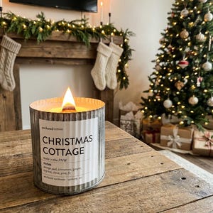 May include: A lit candle in a corrugated metal container with a white label reading Christmas Cottage, made in the PNW, with notes of orange peel, cinnamon, ginger, cypress, clove, pine, and fir, produced by Anchored Northwest in Spokane, WA.