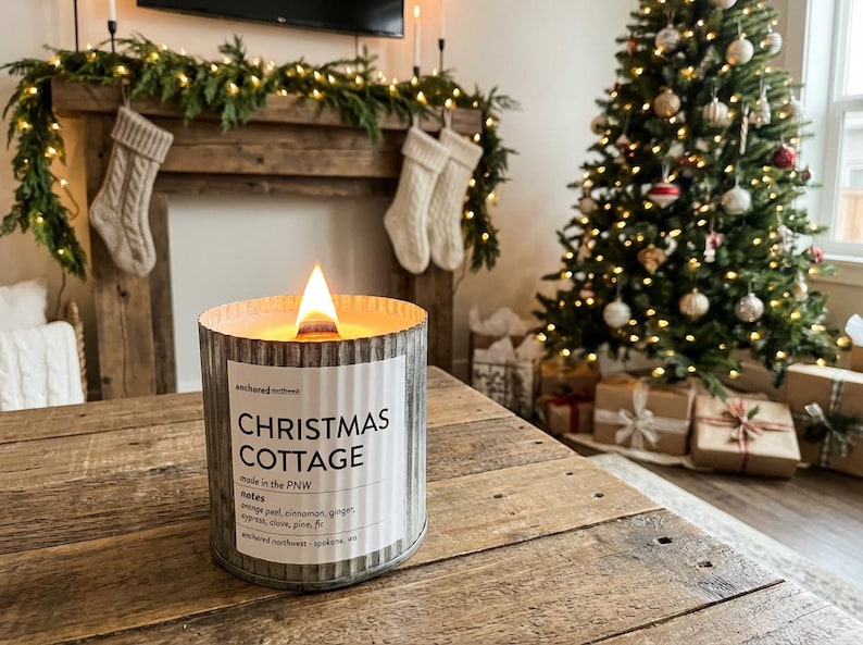 May include: A lit candle in a corrugated metal container with a white label reading Christmas Cottage, made in the PNW, with notes of orange peel, cinnamon, ginger, cypress, clove, pine, and fir, produced by Anchored Northwest in Spokane, WA.