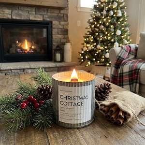 May include: A lit candle in a corrugated metal container with a label reading CHRISTMAS COTTAGE, anchored northwest, made in the PNW, notes orange peel, cinnamon, ginger, cypress, clove, pine, fir, accompanied by pine branches, berries, and cinnamon.