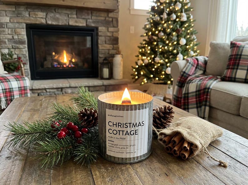 May include: A lit candle in a corrugated metal container with a label reading CHRISTMAS COTTAGE, anchored northwest, made in the PNW, notes orange peel, cinnamon, ginger, cypress, clove, pine, fir, accompanied by pine branches, berries, and cinnamon.