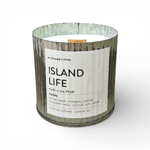 May include: A cylindrical candle in a distressed metal container. The label reads "ISLAND LIFE" with scent notes listed below. The candle has a wooden wick and is from "anchored northwest" in Spokane, WA.