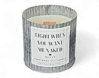 Puede incluir: Una vela cilíndrica en un recipiente de vidrio texturizado de color plateado. La vela tiene un interior de cera blanca y una mecha de madera. Una etiqueta blanca en la parte delantera dice "LIGHT WHEN YOU WANT ME NAKED".