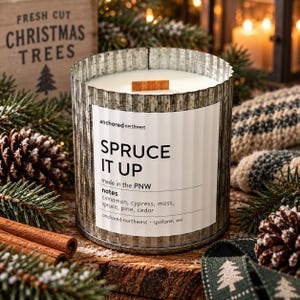 May include: A cylindrical candle in a corrugated galvanized metal container with a white label reading Spruce It Up, anchored northwest, made in the PNW, and notes of cinnamon, cypress, moss, spruce, pine, and cedar with a wooden wick.
