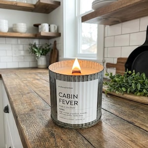 May include: A lit candle in a galvanized metal container with the text "Cabin Fever" on a label. The candle sits on a wooden countertop in a kitchen setting. The candle's scent notes include sage, orange, and grapefruit.