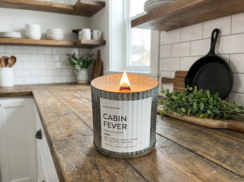 May include: A lit candle in a galvanized metal container with the text "Cabin Fever" on a label. The candle sits on a wooden countertop in a kitchen setting. The candle's scent notes include sage, orange, and grapefruit.