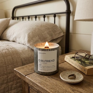 May include: A lit Girlfriend candle in a corrugated metal container sits on a wooden bedside table. The candle has a white label with the word "Girlfriend" and scent notes. A small dish with a ring and a book with glasses are also on the table.