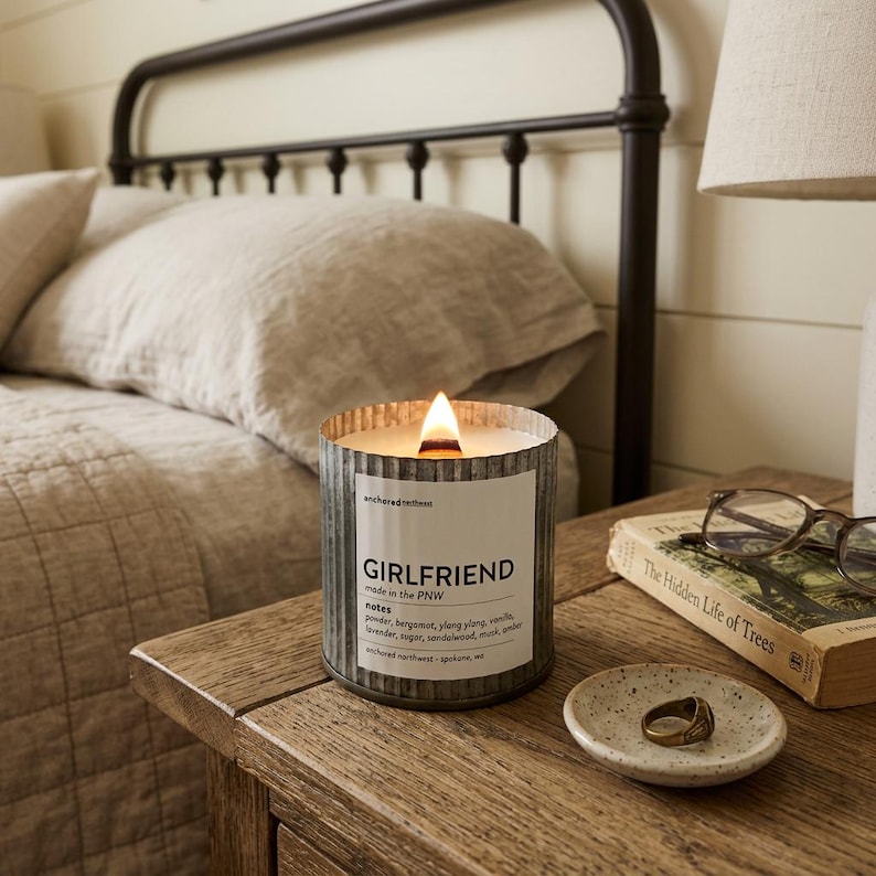 May include: A lit Girlfriend candle in a corrugated metal container sits on a wooden bedside table. The candle has a white label with the word "Girlfriend" and scent notes. A small dish with a ring and a book with glasses are also on the table.
