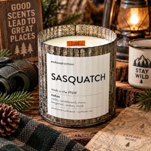 May include: A cylindrical candle in a corrugated metal container with a white label reading SASQUATCH, made in the PNW, and notes of cedar, sandalwood, moss, bergamot, amber, and musk. The candle features a single wooden wick and sits upright.