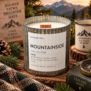Puede incluir: Una vela arom&aacute;tica en un recipiente de metal galvanizado corrugado con una etiqueta blanca que dice MOUNTAINSIDE, anchored northwest, made in the PNW, y notas de s&aacute;ndalo, cilantro, baya de enebro, naranja, romero, jengibre y vainilla.