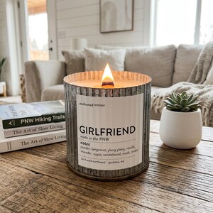 May include: A burning candle in a corrugated metal container with a label reading "GIRLFRIEND." The candle sits on a wooden table, beside a small succulent in a white pot and books titled "PNW Hiking Trails" and "The Art of Slow Living."