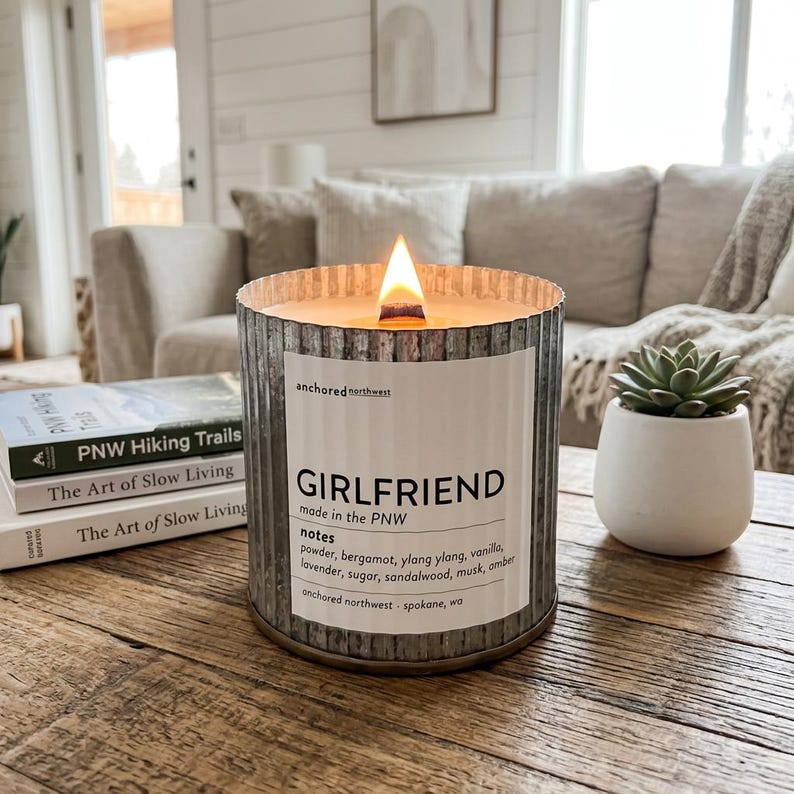 May include: A burning candle in a corrugated metal container with a label reading "GIRLFRIEND." The candle sits on a wooden table, beside a small succulent in a white pot and books titled "PNW Hiking Trails" and "The Art of Slow Living."