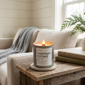 May include: A lit candle in a cylindrical, corrugated galvanized metal container with a white label reading Christmas Cottage, made in the PNW, notes: orange peel, cinnamon, ginger, cypress, clove, pine, fir, anchored northwest, spokane, wa.