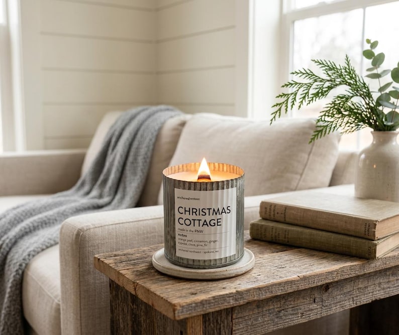 May include: A lit candle in a cylindrical, corrugated galvanized metal container with a white label reading Christmas Cottage, made in the PNW, notes: orange peel, cinnamon, ginger, cypress, clove, pine, fir, anchored northwest, spokane, wa.