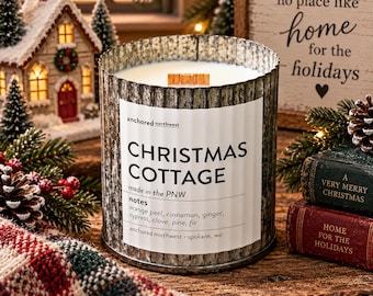 Puede incluir: Una vela cilíndrica en un recipiente de metal corrugado con una etiqueta blanca que dice Christmas Cottage, made in the PNW, con notas de cáscara de naranja, canela, jengibre, ciprés, clavo, pino y abeto. Incluye una mecha de madera.