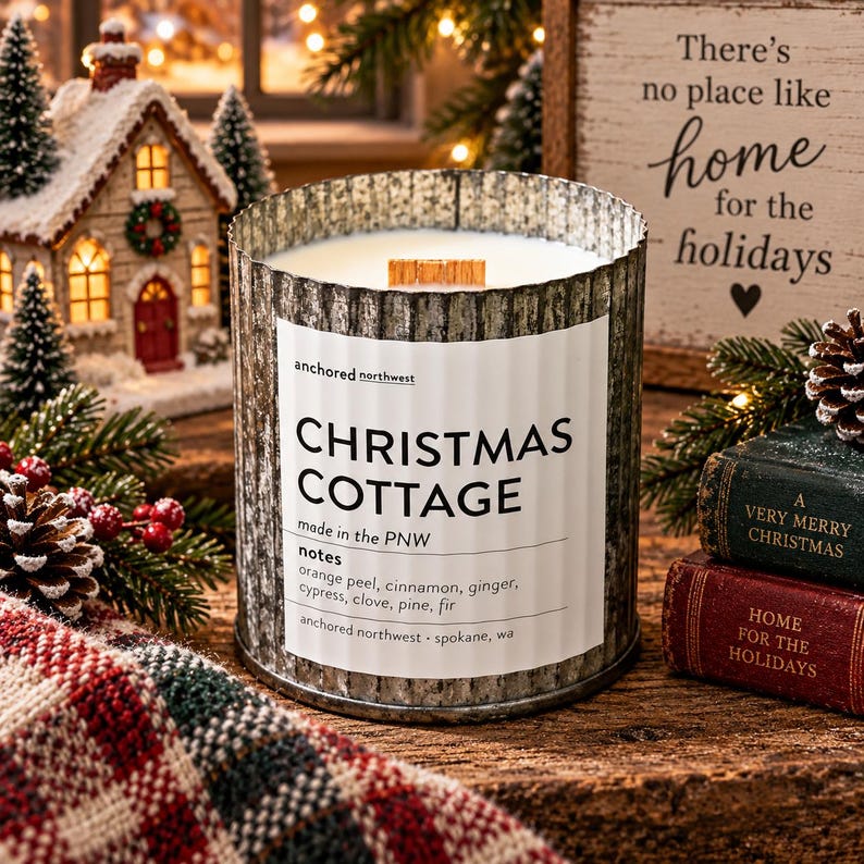 May include: A cylindrical candle in a corrugated metal container with a white label reading Christmas Cottage, made in the PNW, with notes of orange peel, cinnamon, ginger, cypress, clove, pine, and fir. It features a wooden wick.