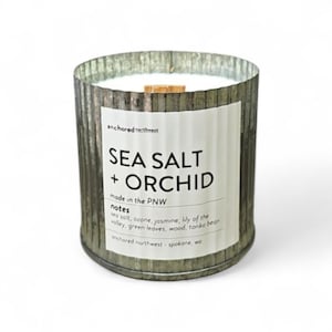 May include: A cylindrical candle in a galvanized metal container. The label reads "SEA SALT + ORCHID" with additional text. The candle has a wooden wick and is filled with white wax. The product is from "anchored northwest" in Spokane, WA.