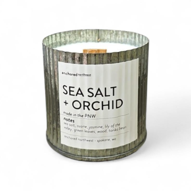 May include: A cylindrical candle in a galvanized metal container. The label reads "SEA SALT + ORCHID" with additional text. The candle has a wooden wick and is filled with white wax. The product is from "anchored northwest" in Spokane, WA.