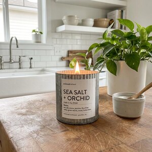 May include: A lit candle in a galvanized metal container with the text "SEA SALT + ORCHID" on a wooden table. A small ceramic bowl with a wooden spoon and a potted plant are also on the table. The candle is in a kitchen setting.