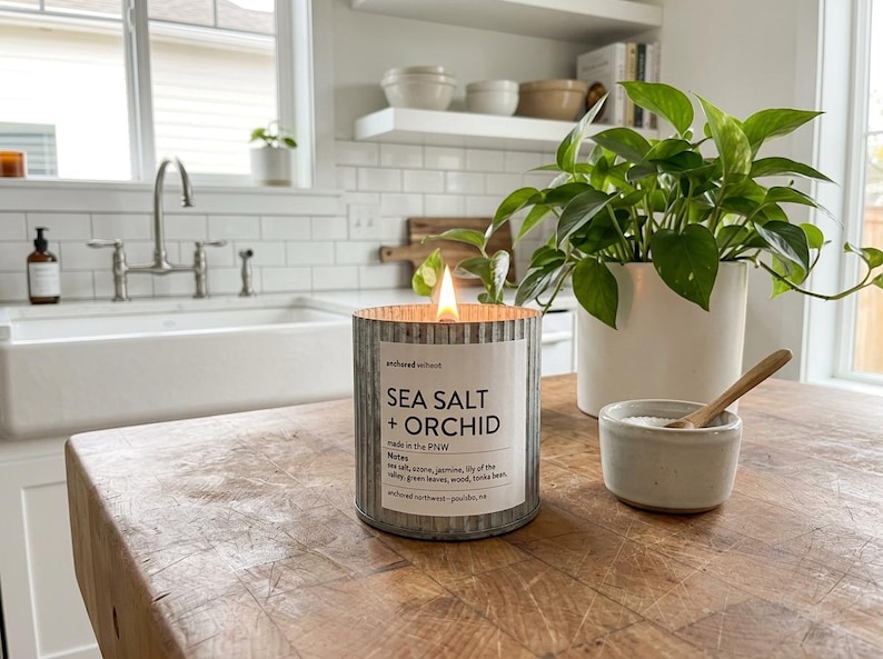 May include: A lit candle in a galvanized metal container with the text "SEA SALT + ORCHID" on a wooden table. A small ceramic bowl with a wooden spoon and a potted plant are also on the table. The candle is in a kitchen setting.