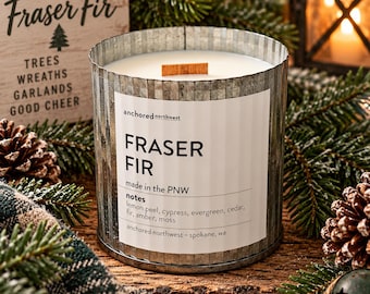 Może przedstawiać: Cylindryczna świeca w karbowanym, ocynkowanym metalowym pojemniku z białą etykietą z napisem Anchored Northwest Fraser Fir, made in the PNW, z nutami skórki cytryny, cyprysu, wiecznie zielonych roślin, cedru, jodły, bursztynu i mchu oraz drewnianym knotem.