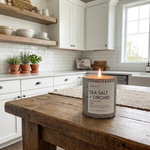 May include: A lit candle in a galvanized metal container with the text "SEA SALT + ORCHID" sits on a rustic wooden table. The candle is in a kitchen with white cabinets, a white sink, and a window.