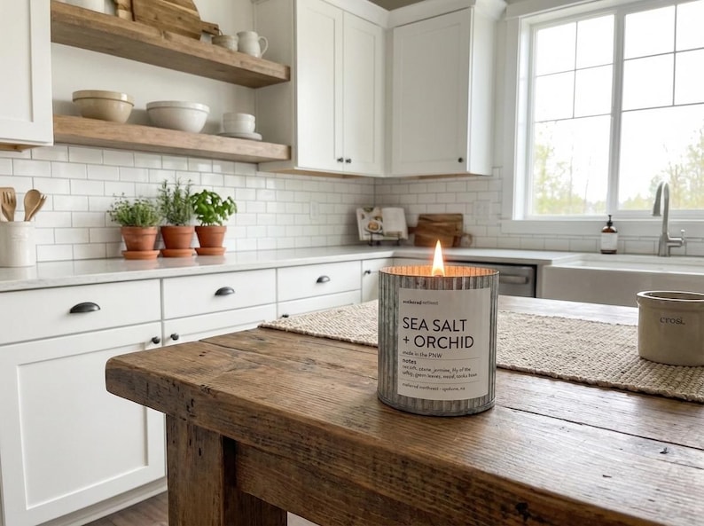May include: A lit candle in a galvanized metal container with the text "SEA SALT + ORCHID" sits on a rustic wooden table. The candle is in a kitchen with white cabinets, a white sink, and a window.