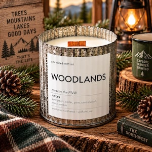 May include: A cylindrical candle in a corrugated metal container with a white label reading WOODLANDS, made in the PNW, with notes of evergreen, cedar, pine, sandalwood, moss, and amber. The candle features a single rectangular wooden wick.