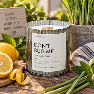 May include: A cylindrical candle in a corrugated galvanized metal container with a white label reading DON'T BUG ME, made in the PNW, notes lemon peel, camphor, lemongrass, citronella, powder, anchored northwest - spokane, wa, and a wooden wick.