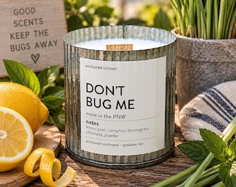 May include: A cylindrical candle in a corrugated galvanized metal container with a white label reading DON'T BUG ME, made in the PNW, notes lemon peel, camphor, lemongrass, citronella, powder, anchored northwest - spokane, wa, and a wooden wick.