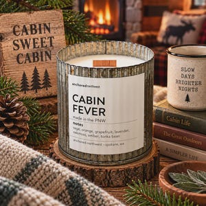 May include: A cylindrical candle in a corrugated metal container with a white label reading Cabin Fever, anchored northwest, made in the PNW, and a list of scent notes including sage, orange, grapefruit, lavender, oakmoss, amber, and tonka bean.